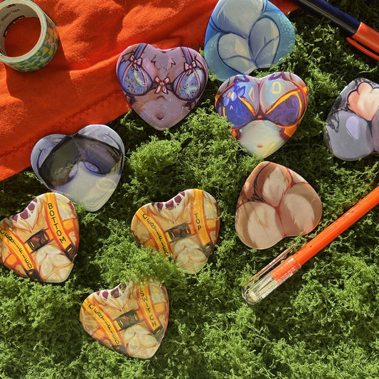 A styled flat-lay of several heart-shaped painted stones or pendants arranged on bright green moss, with an orange fabric, washi tape, and orange pen as props.