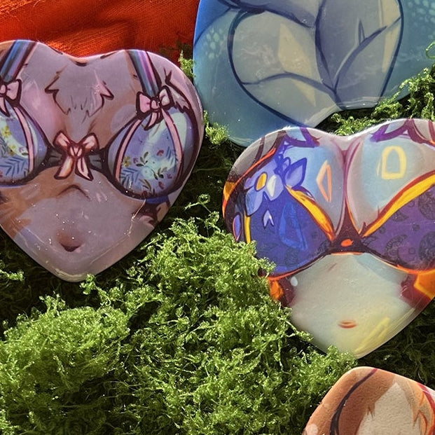 A styled flat-lay of several heart-shaped painted stones or pendants arranged on bright green moss, with an orange fabric, washi tape, and orange pen as props.