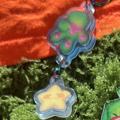 A collection of colorful cartoon acrylic keychains and charms displayed on bright green moss, with an orange fabric background.