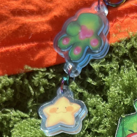 A collection of colorful cartoon acrylic keychains and charms displayed on bright green moss, with an orange fabric background.